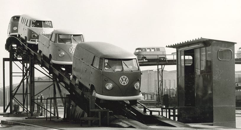 Trzy samochody marki Volkswagen Transporter zjeżdżają po metalowej rampie w fabryce, a w tle widać inne pojazdy.