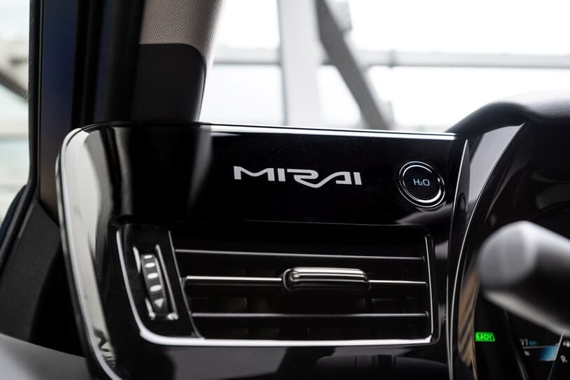 Kokpit Toyoty Mirai z napisem „MIRAI”, przyciskiem „H2O” i fragmentem kierownicy.
