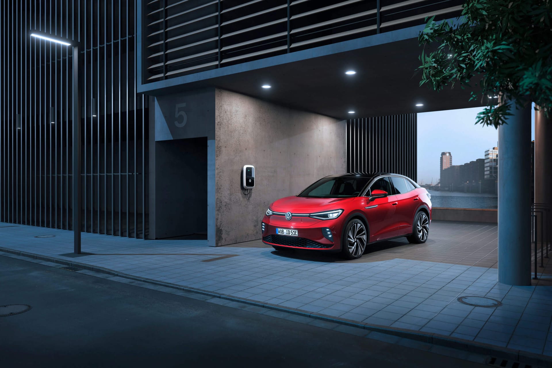 Czerwony Volkswagen ID.5 ładuje się o zmierzchu w miejskim garażu pod dachem, a w tle widać rzekę i budynki.