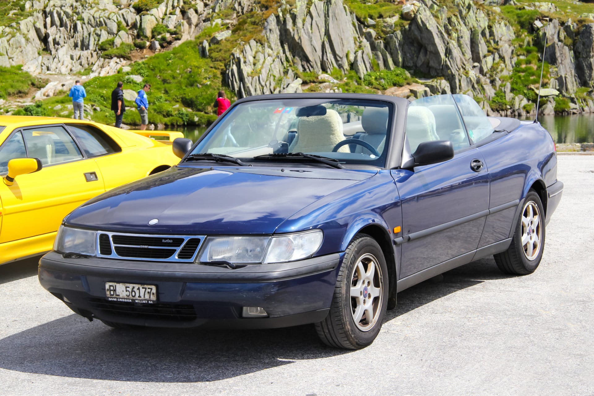 Niebieski Saab 900 kabriolet stoi na bruku, a w tle widać skały i zieloną trawę.