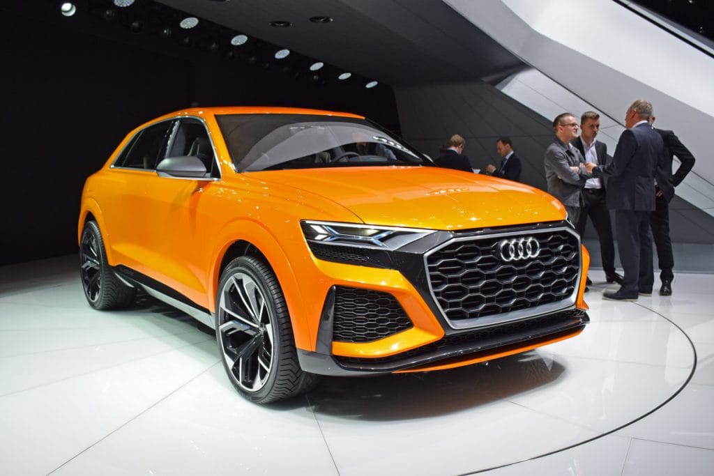 Pomarańczowy SUV marki Audi na platformie, a w tle czterech mężczyzn w garniturach na targach.