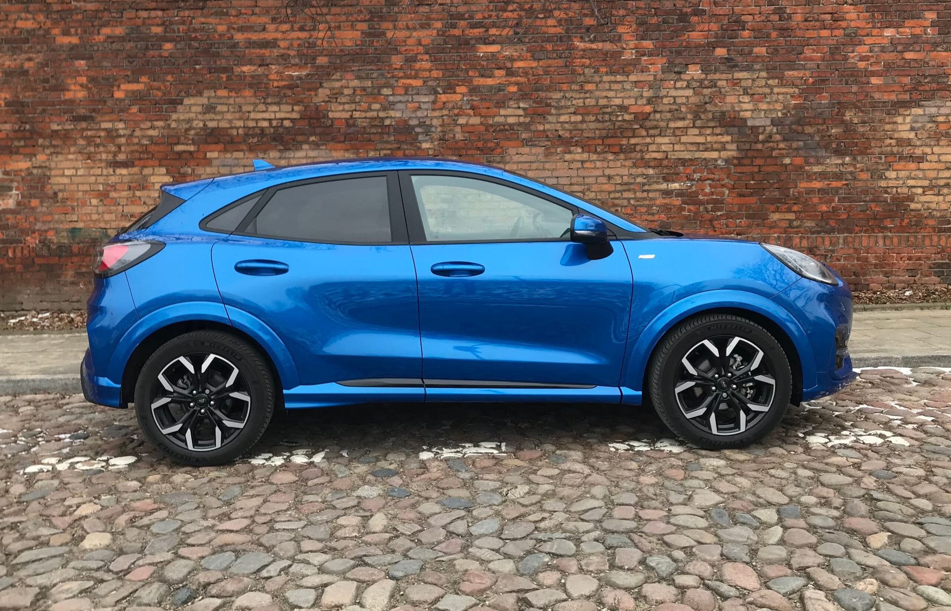 Niebieski Ford Puma stoi na brukowanej ulicy przed czerwoną ceglaną ścianą.