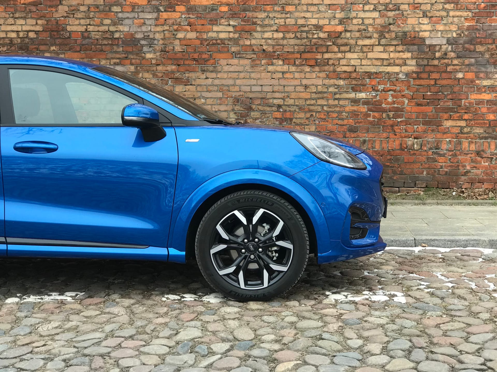 Niebieski Ford Puma zaparkowany przodem na brukowanej ulicy przy starej ceglanej ścianie.