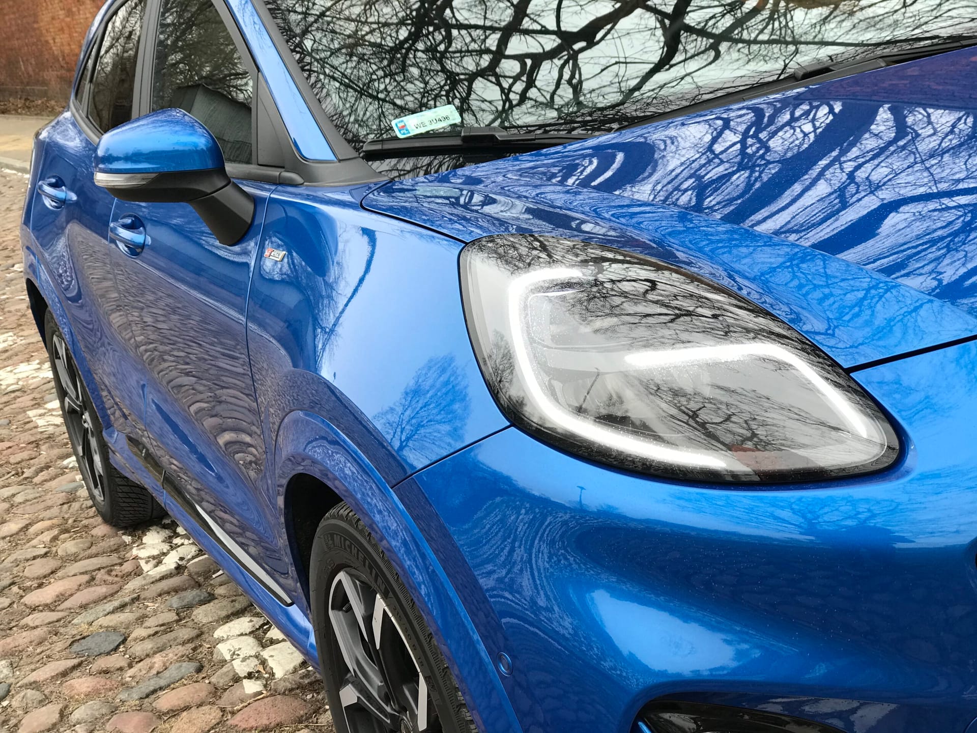 Niebieski Ford Puma widziany z przodu na bruku, odbicia gałęzi na masce i przedniej szybie.