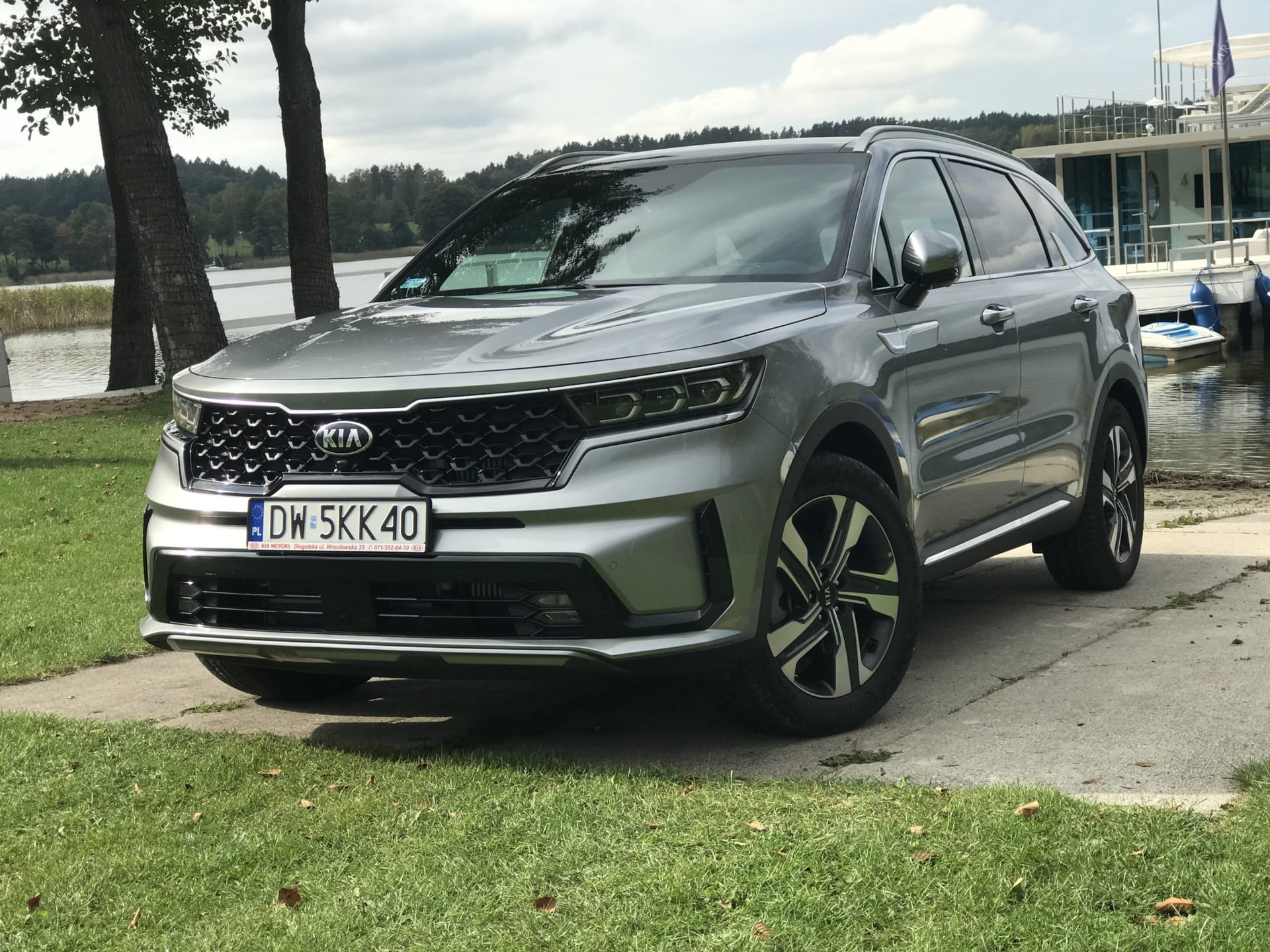 Srebrny Kia Sorento zaparkowany nad jeziorem, w tle drzewa i zacumowana łódź.