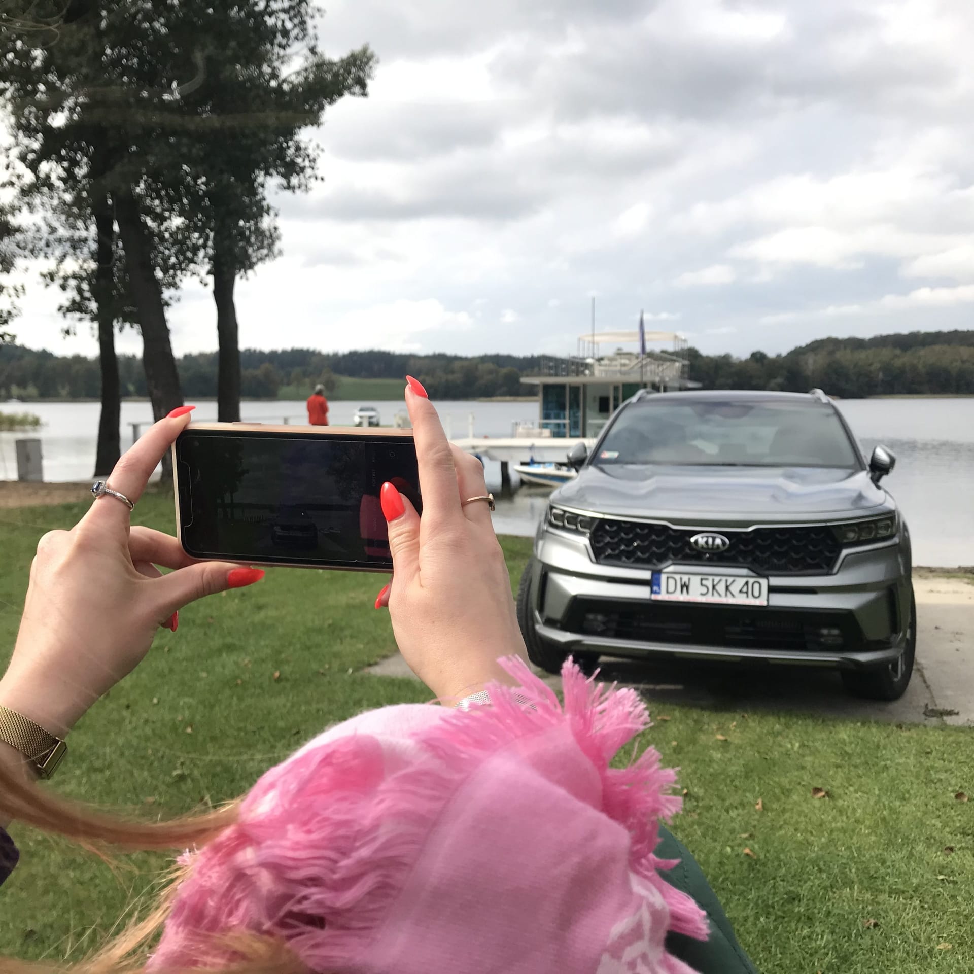 Osoba w różowym szaliku fotografuje zaparkowanego nad jeziorem Kia Sorento, drzewa i łódki.