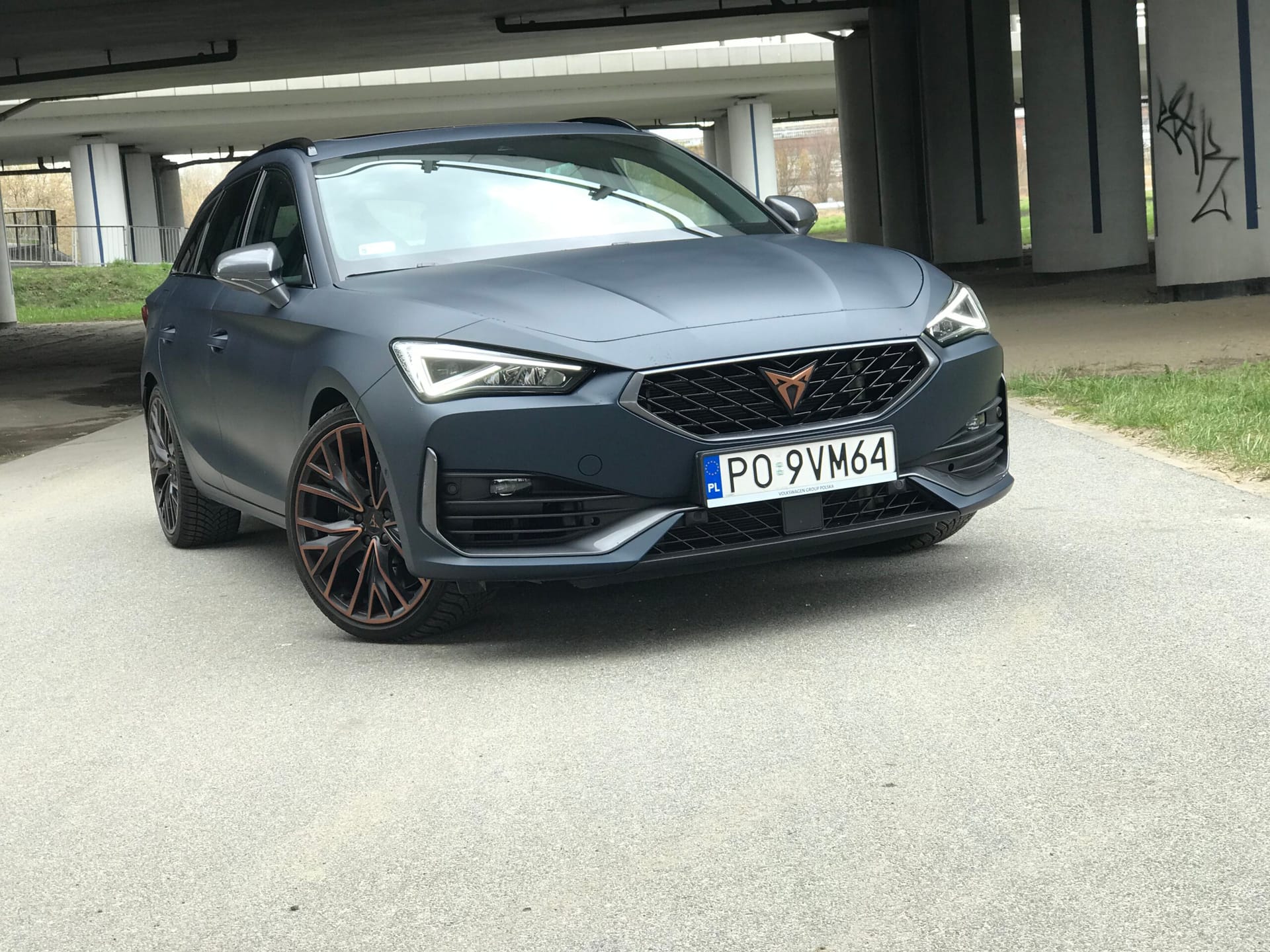 Matowy niebieski Cupra Leon ST z polskimi tablicami rejestracyjnymi stoi na asfalcie pod mostem.