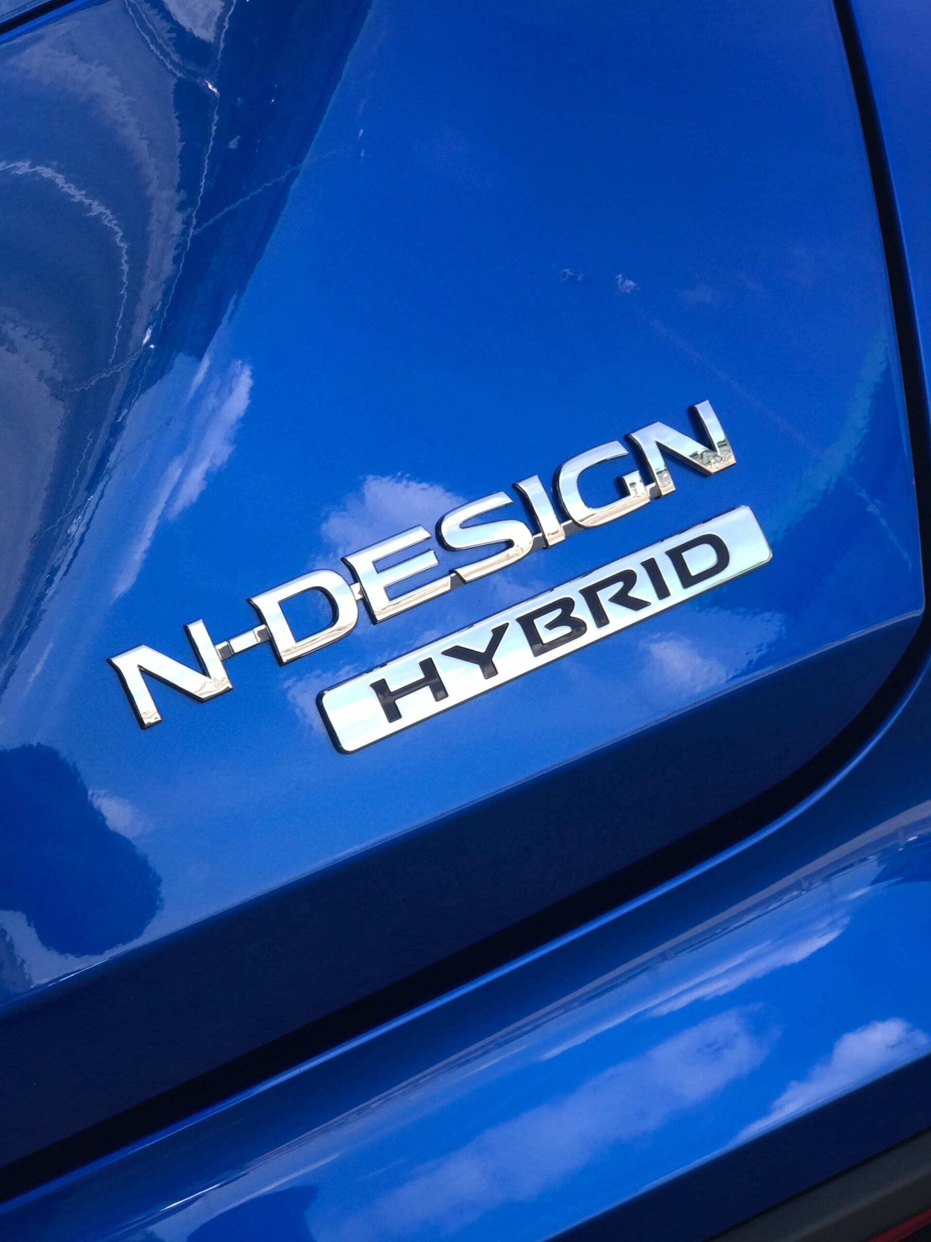Zbliżenie na niebieskiego Nissana Juke Hybrid z chromowanymi emblematami N-DESIGN i HYBRID.
