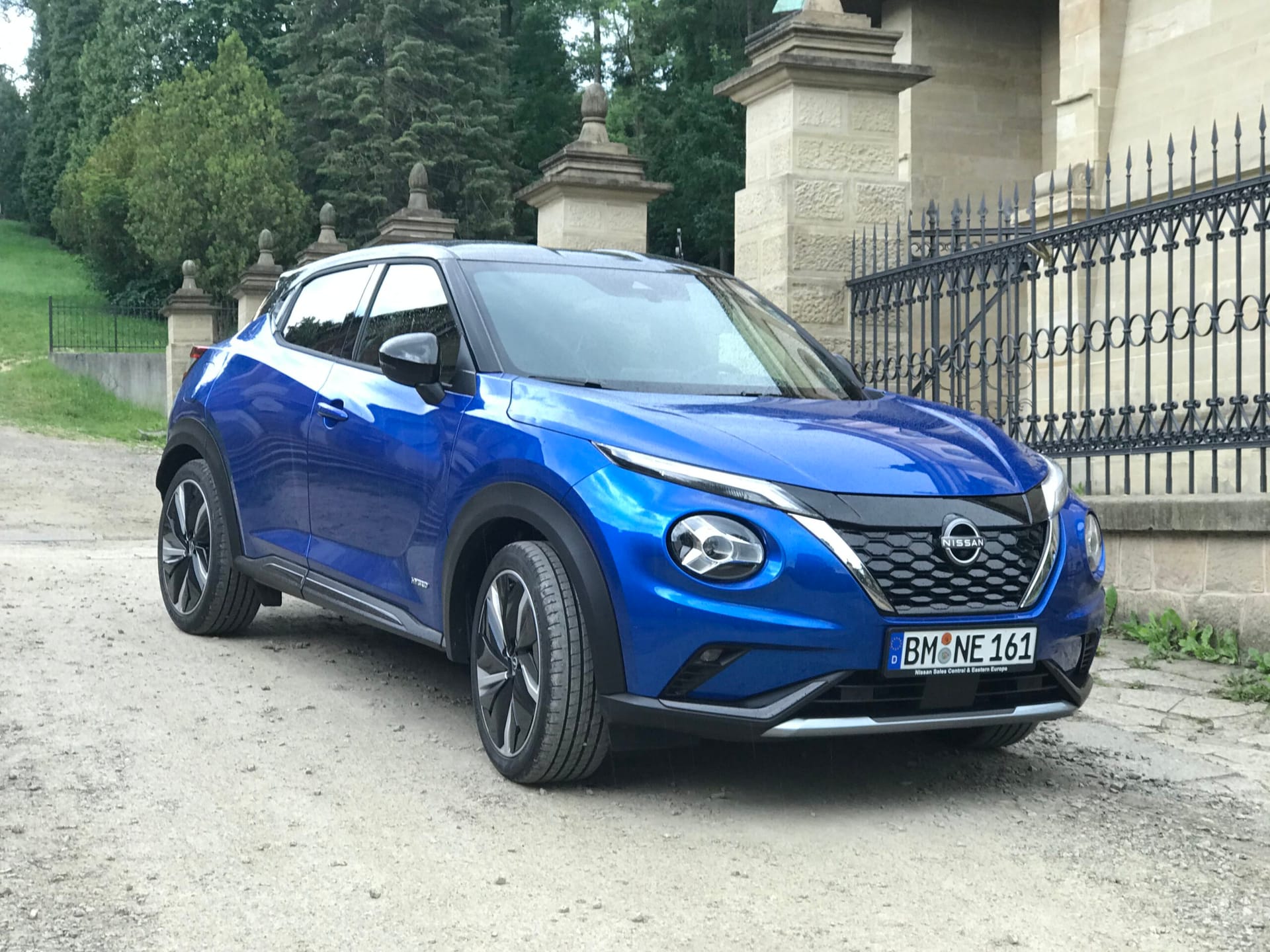 Niebieski Nissan Juke Hybrid stoi na polnej drodze przy czarnym ogrodzeniu i kamiennym budynku.