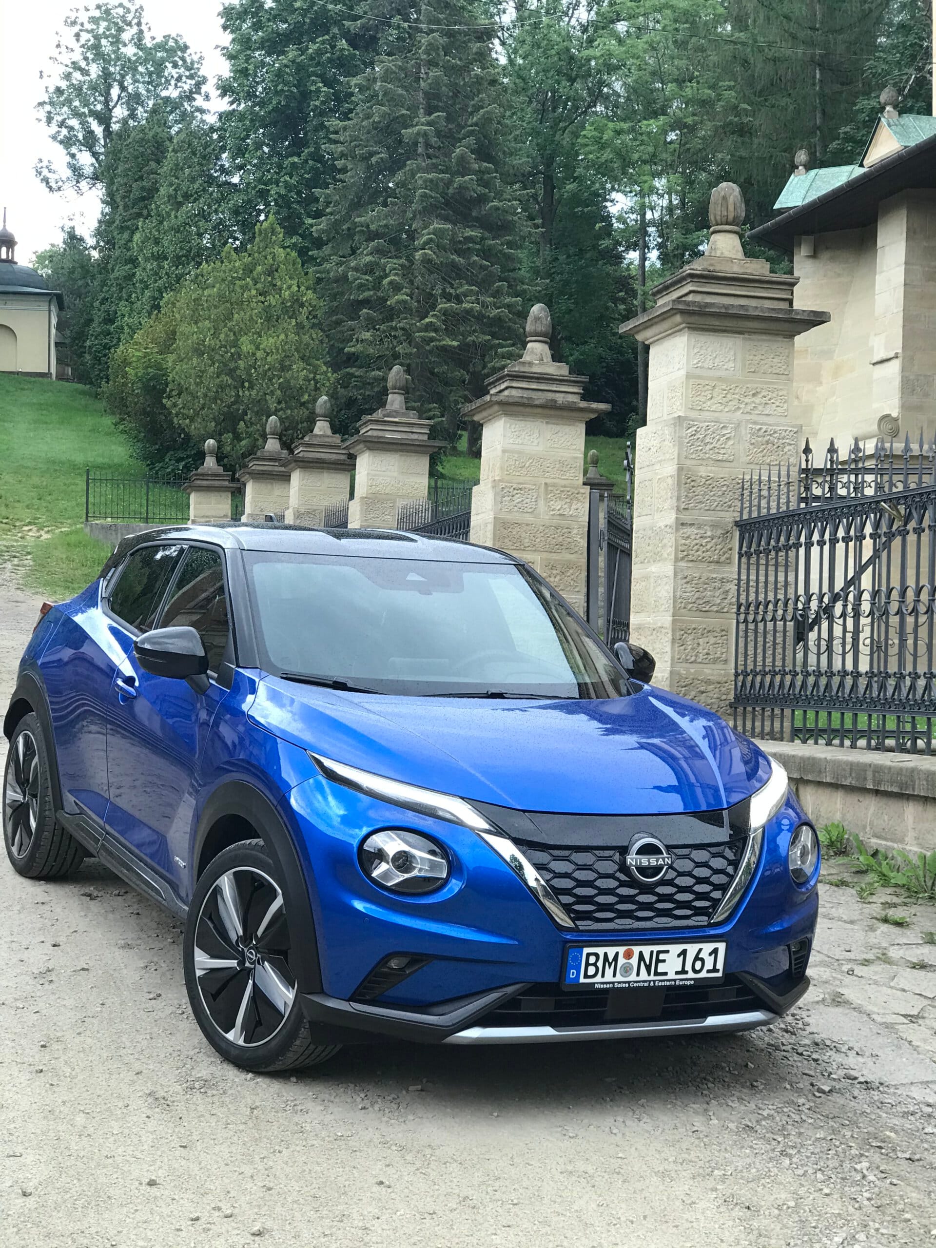 Niebieski Nissan Juke Hybrid stoi na polnej drodze obok kamiennego muru i zieleni.