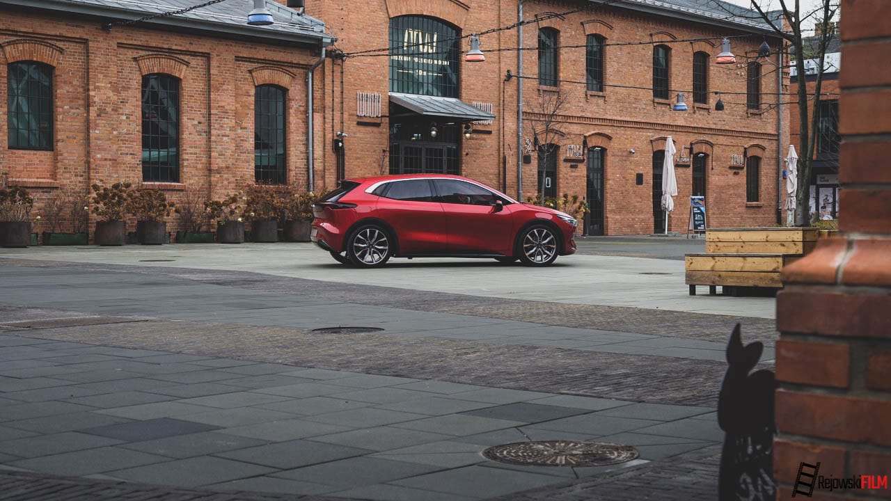 Czerwony hatchback, prawdopodobnie Izera, zaparkowany na miejskim dziedzińcu otoczonym ceglanymi budynkami.