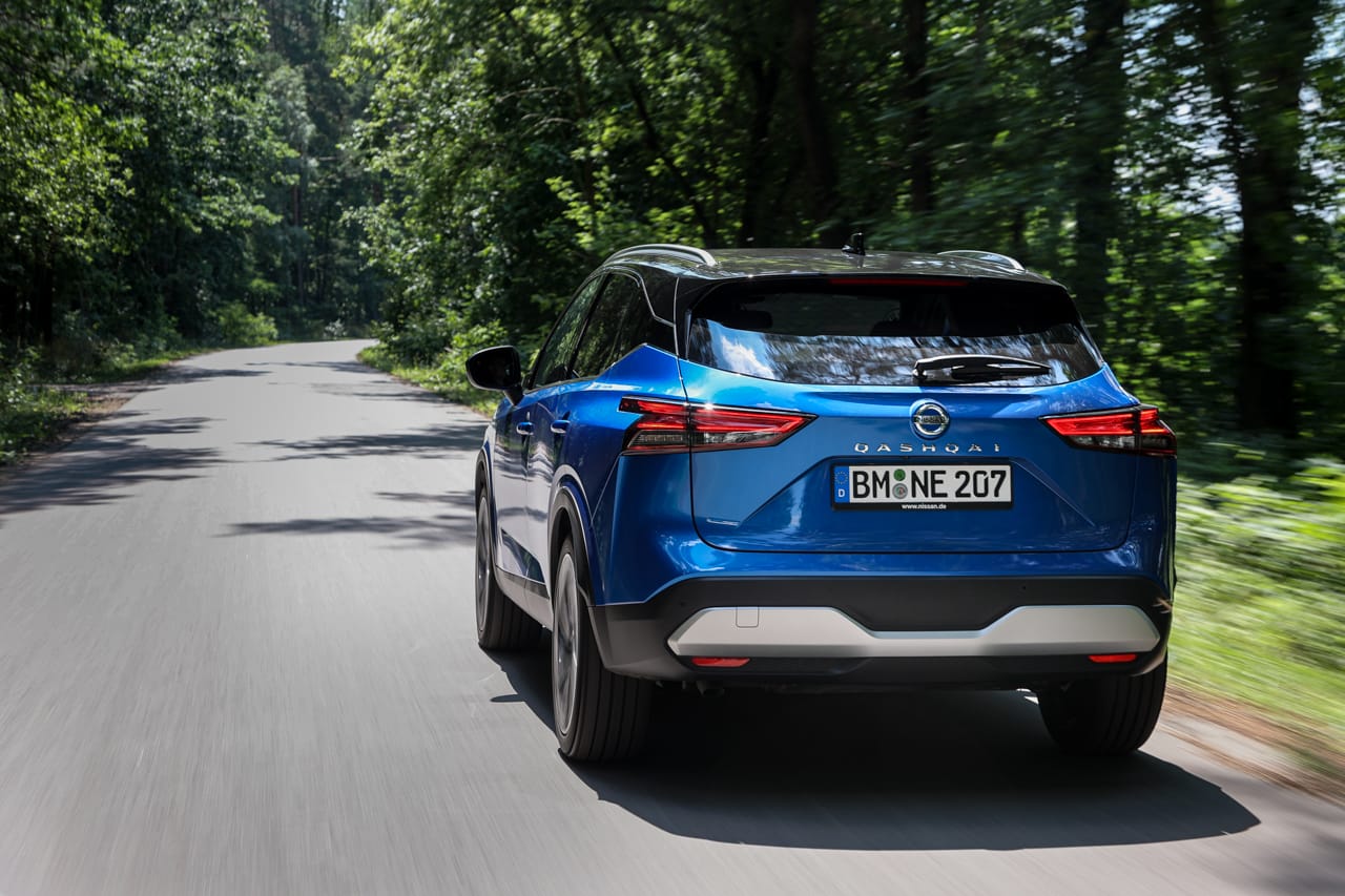 Niebieski Nissan Qashqai jedzie krętą drogą przez zielony las w słoneczny dzień.