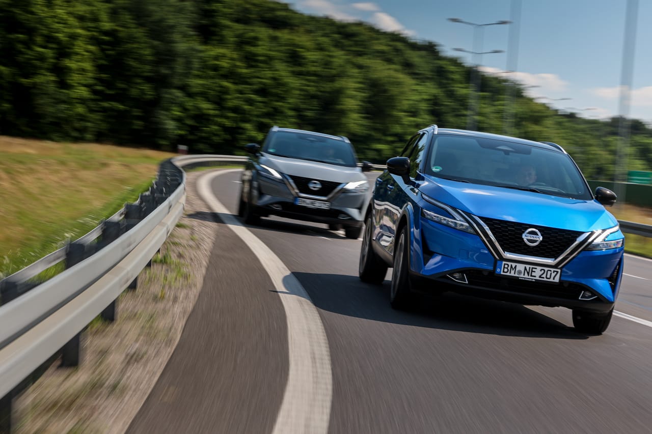 Dwa niebieskie SUV-y, w tym Nissan Qashqai, jadą krętą drogą na tle drzew.