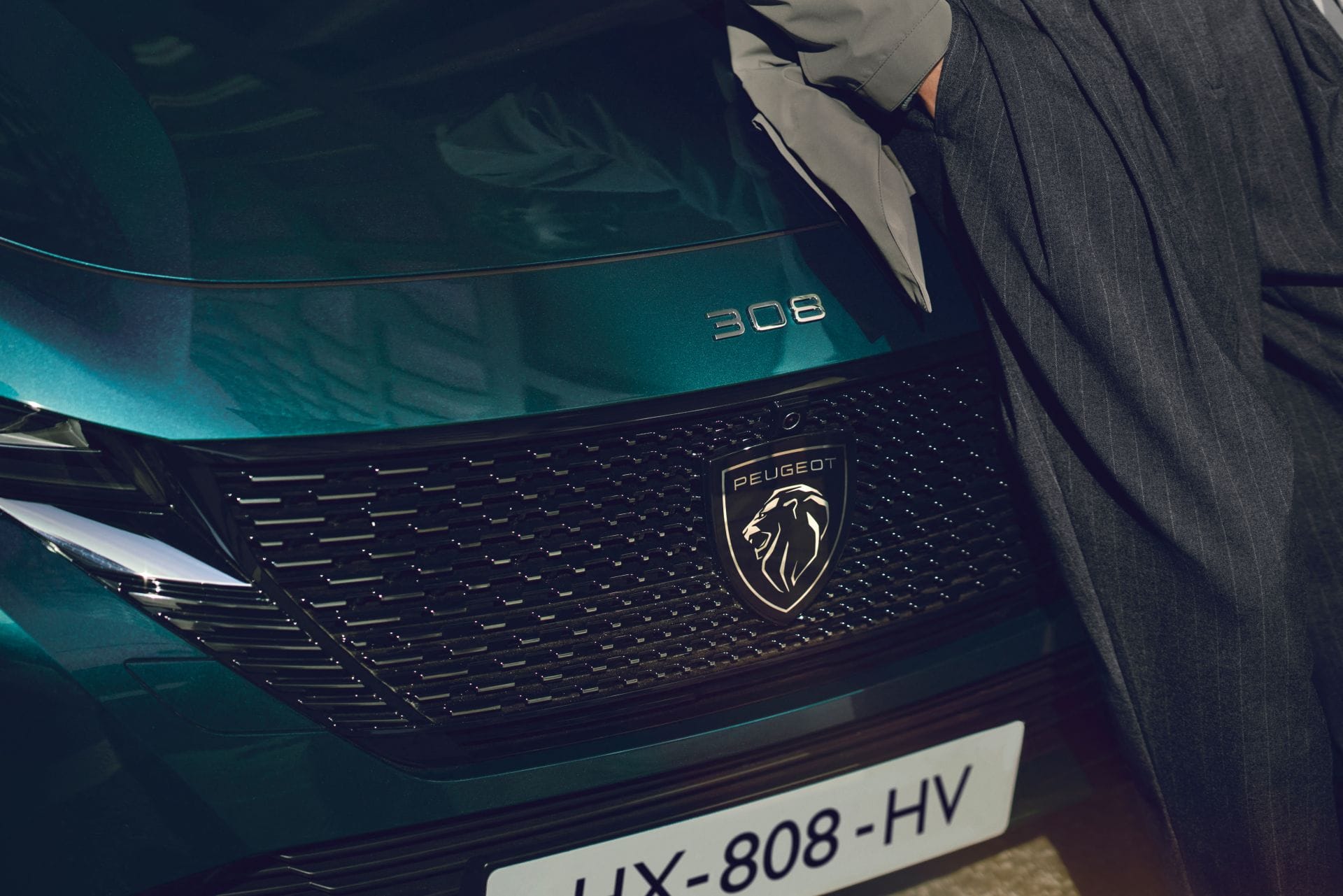 Zbliżenie na grill i emblemat Peugeota 308 SW, obok fragmentu sylwetki osoby i tablicy.