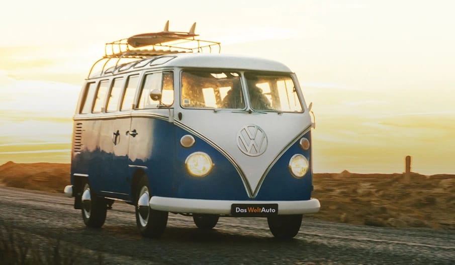 Niebiesko-biały Volkswagen Transporter z deską surfingową jedzie o zachodzie słońca.