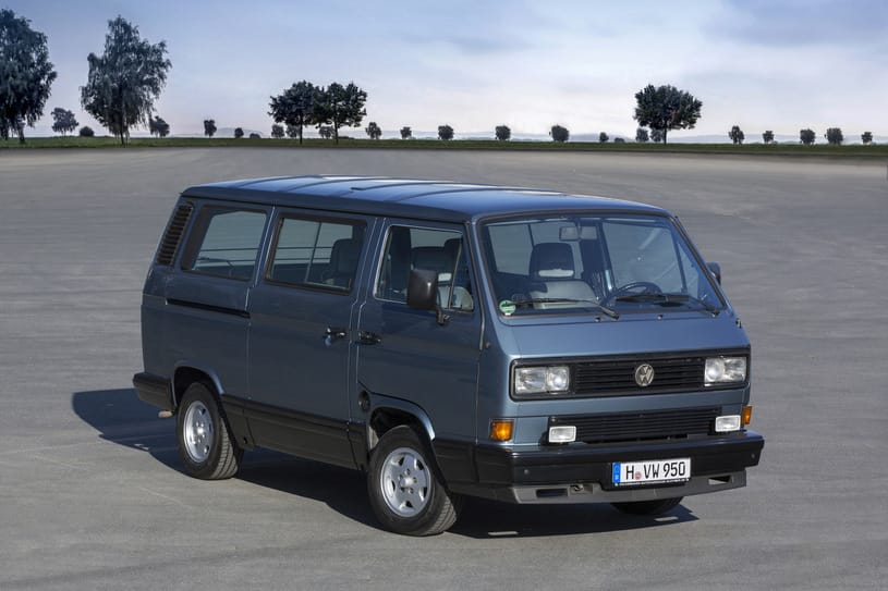 Niebieski Volkswagen Transporter Vanagon stoi na otwartym placu, a w tle widać drzewa i chmury.