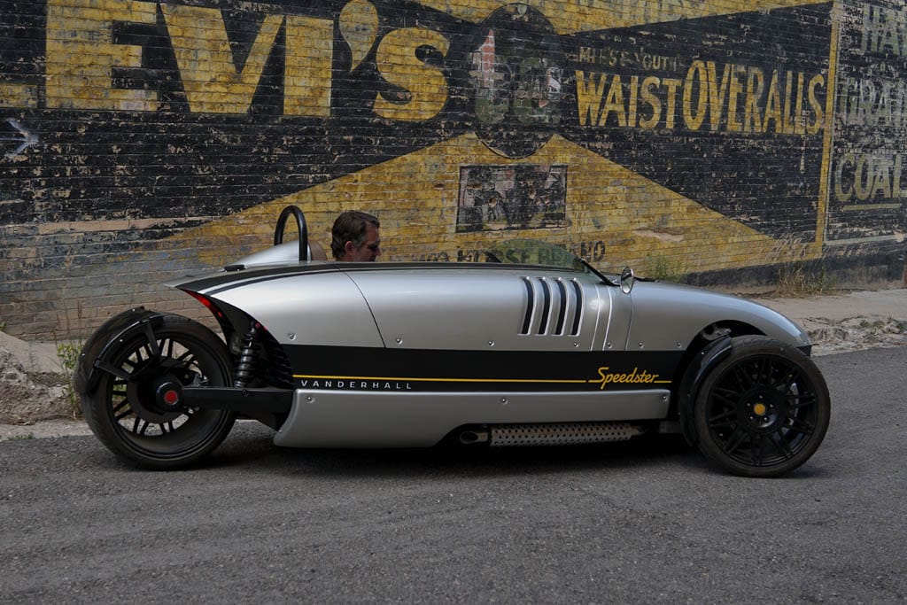 Srebrny Vanderhall Speedster na trzech kołach stoi przy wyblakłej ścianie z reklamą Levi’s.