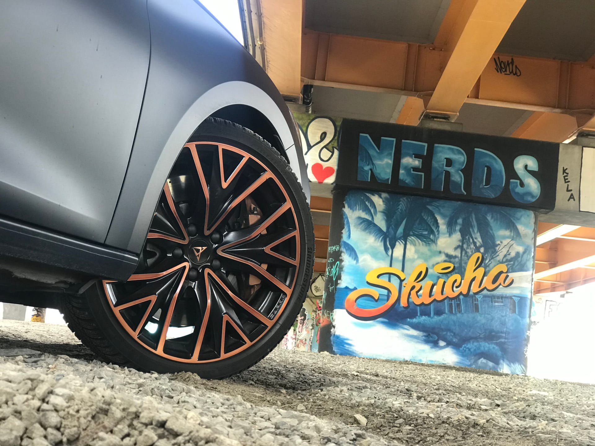 Cupra Leon ST z pomarańczowymi akcentami na żwirze, a w tle graffiti NERDS i Skucha.