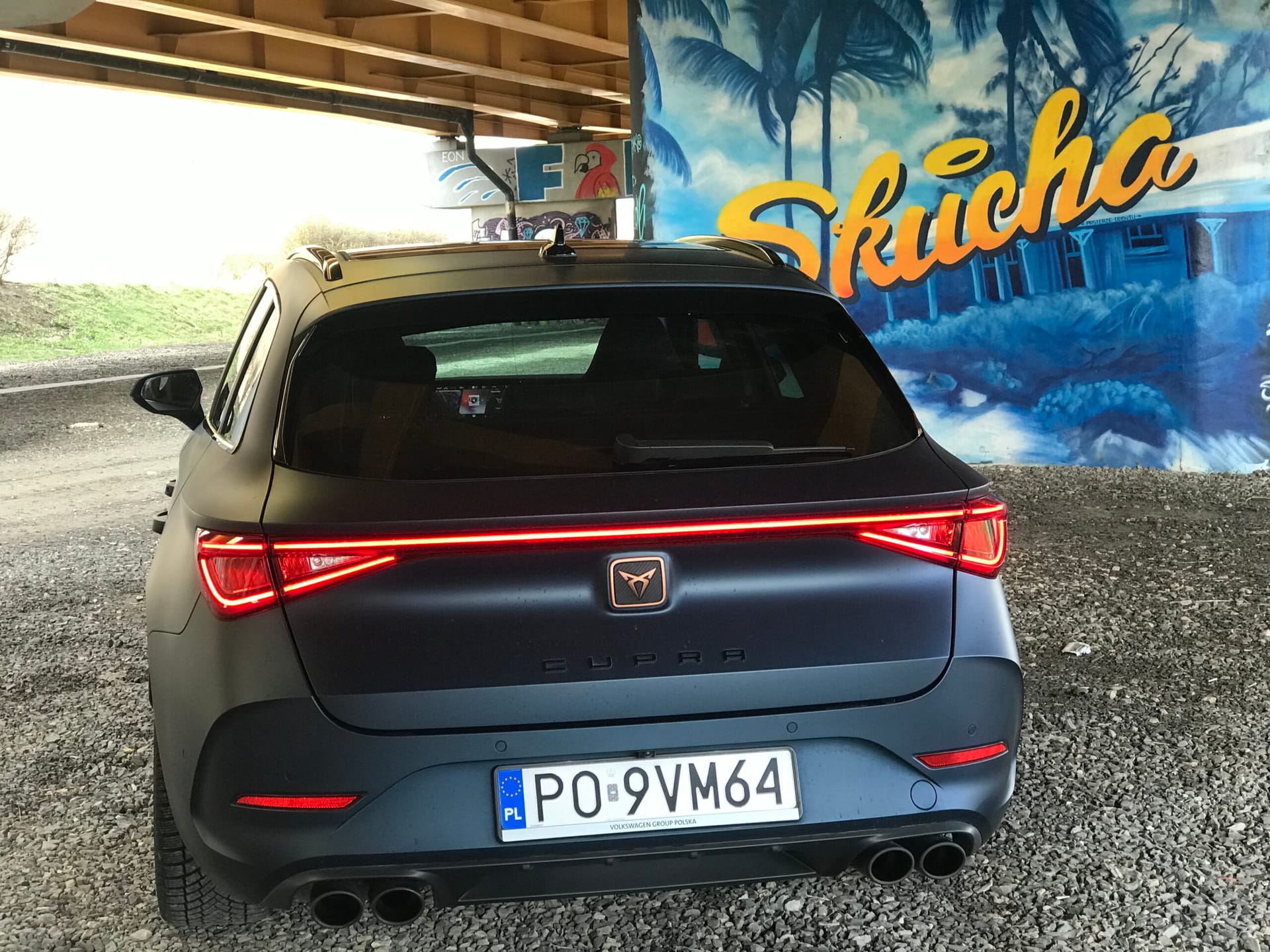 Czarny Cupra Leon ST z polskimi tablicami rejestracyjnymi stoi pod mostem obok kolorowego muralu przedstawiającego palmy i niebieski dom.