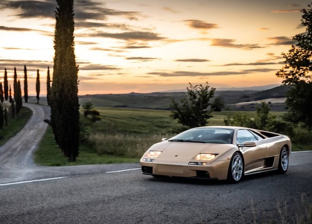 Złote Lamborghini Diablo jedzie wiejską drogą o zachodzie słońca, mijając drzewo i wzgórze.