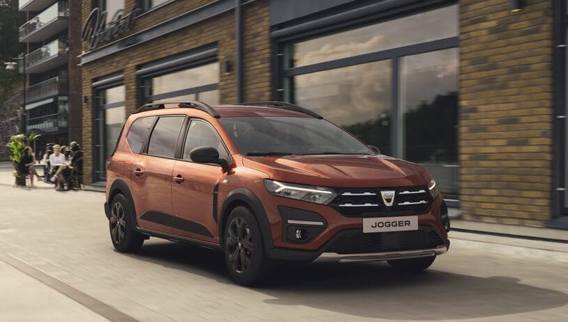 Czerwony Dacia Jogger jedzie ulicą obok ceglanego budynku z dużymi oknami.