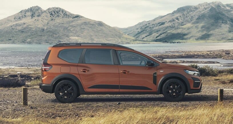 Pomarańczowy SUV Dacia Jogger na żwirowej drodze nad jeziorem, w tle góry i chmury.