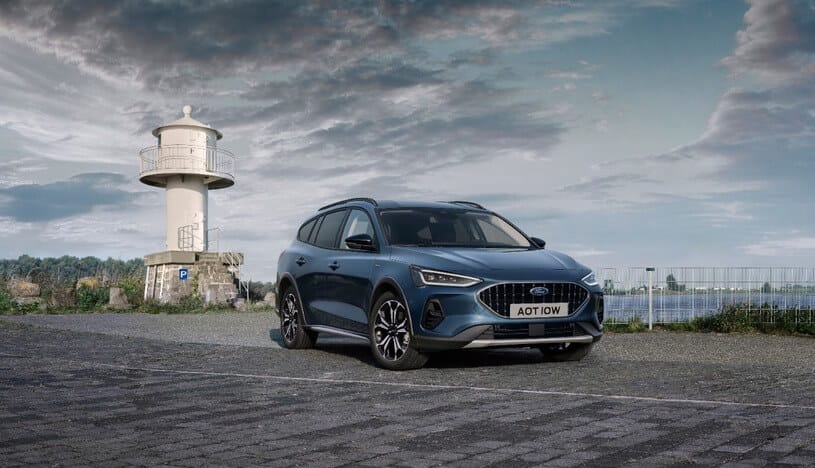Niebieski SUV marki Ford Focus stoi na asfalcie obok białej latarni, a w tle widać wodę i chmury.