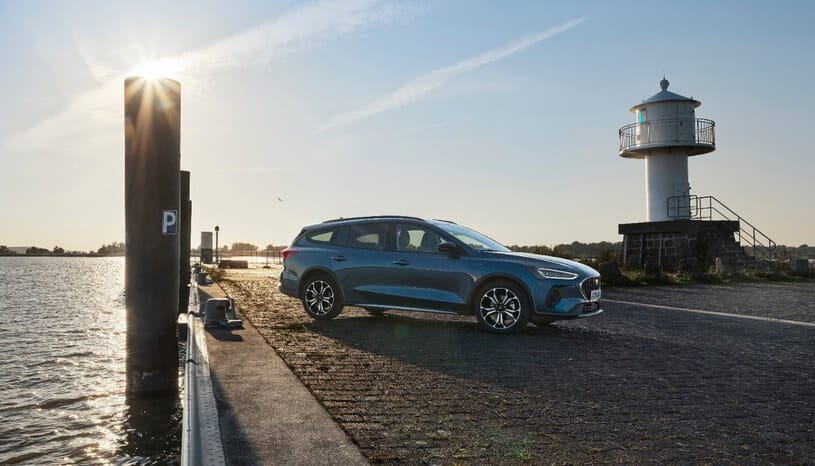 Niebieski Ford Focus kombi stoi nad wodą przy białej latarni o zachodzie słońca.