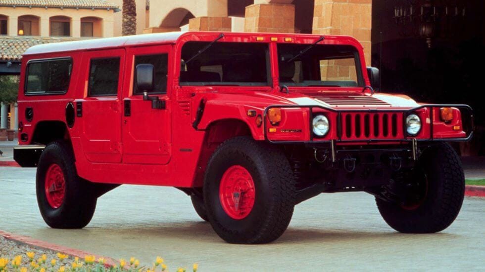 Czerwony Hummer H1 stoi na podjeździe przed budynkiem z łukowatymi oknami i beżowymi ścianami.