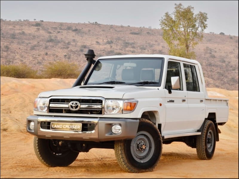 Biały pickup Land Cruiser stoi na piaszczystym terenie wśród pagórków i rzadkich drzew.