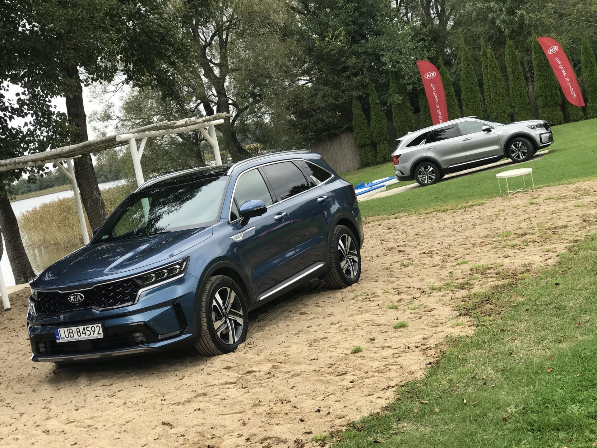Dwa SUV-y marki Kia, w tym Sorento, stoją na piasku nad jeziorem, a obok nich wiszą czerwone banery.