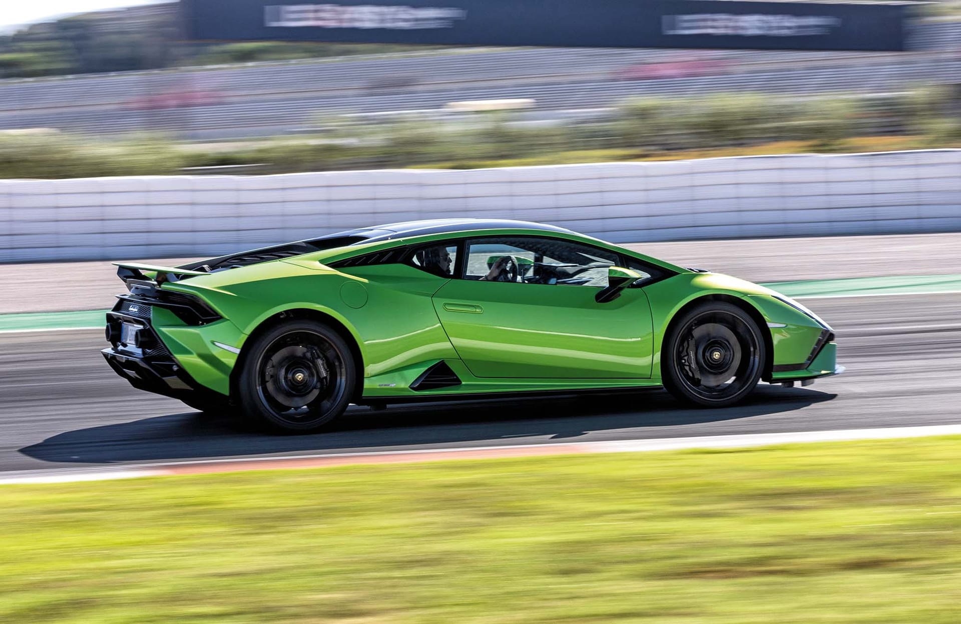 Zielony Lamborghini Huracan Tecnica pędzi po torze, a w tle widać trawę i barierę.