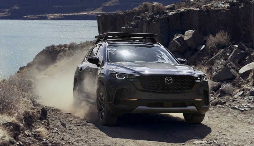 Czarna Mazda CX-50 jedzie po zakurzonej drodze w pobliżu skał i jeziora.
