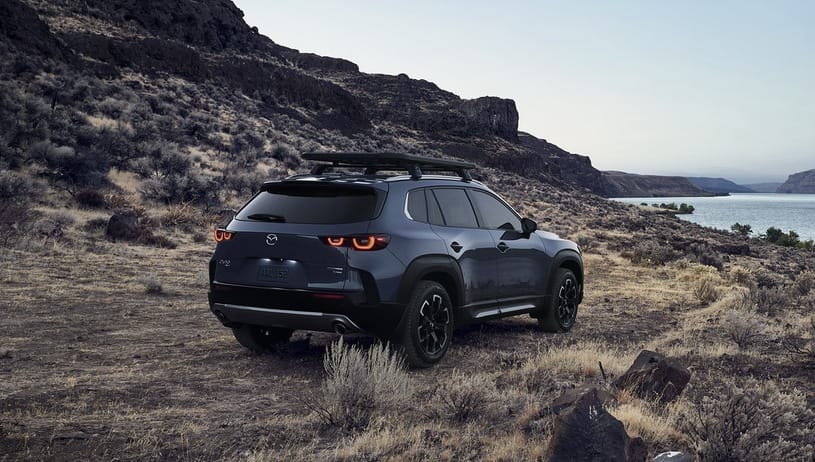 Granatowa Mazda CX-50 stoi na kamienistym terenie porośniętym trawą nad wodą, pod chmurami.