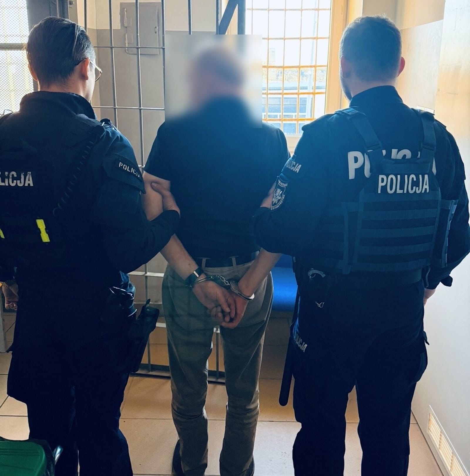 Dwóch policjantów prowadzi skuty kajdankami mężczyznę podejrzanego o złamanie zakazu prowadzenia pojazdów.