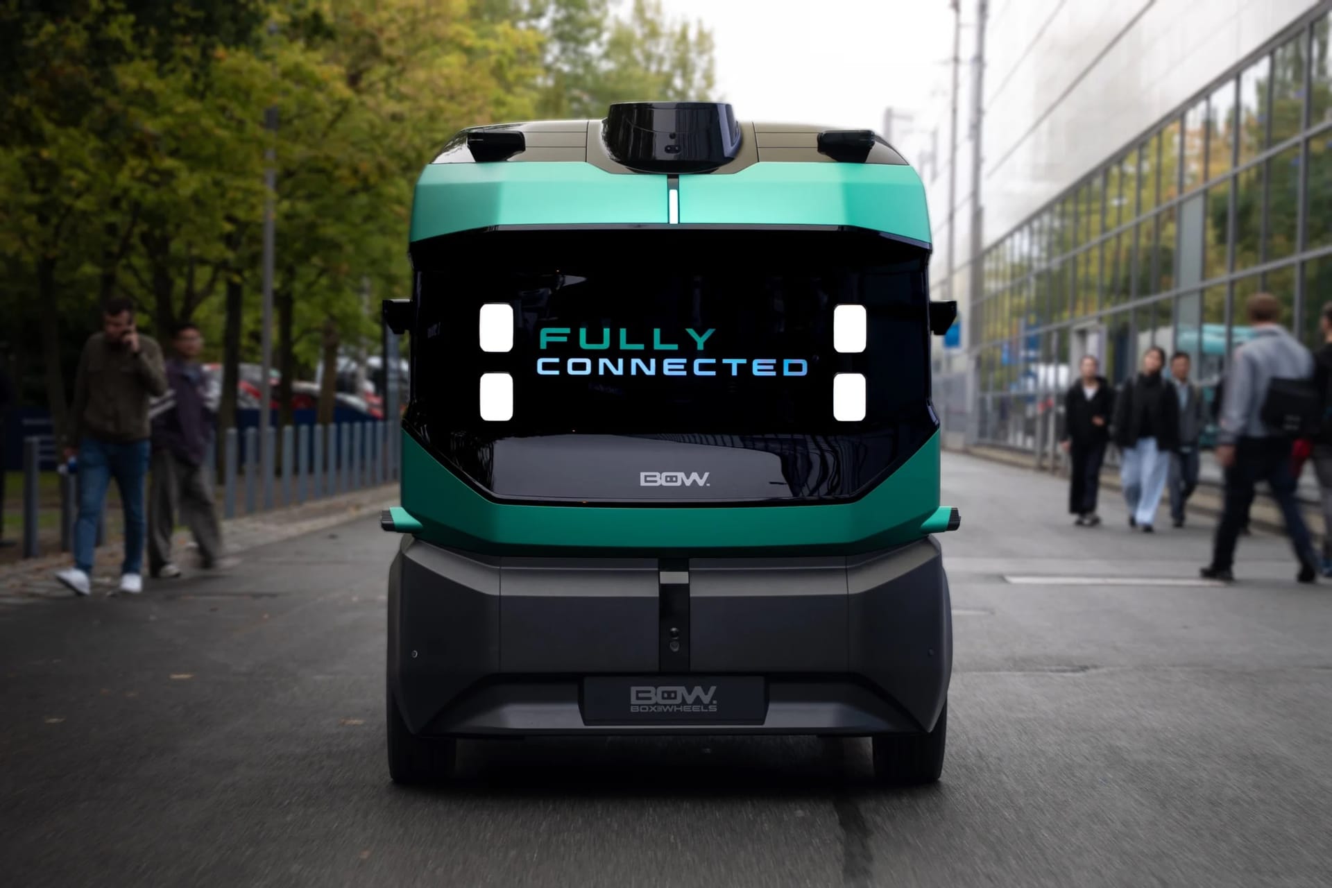 Zielony pojazd autonomiczny z napisem „FULLY CONNECTED” jedzie ulicą, a w tle widać przechodniów.