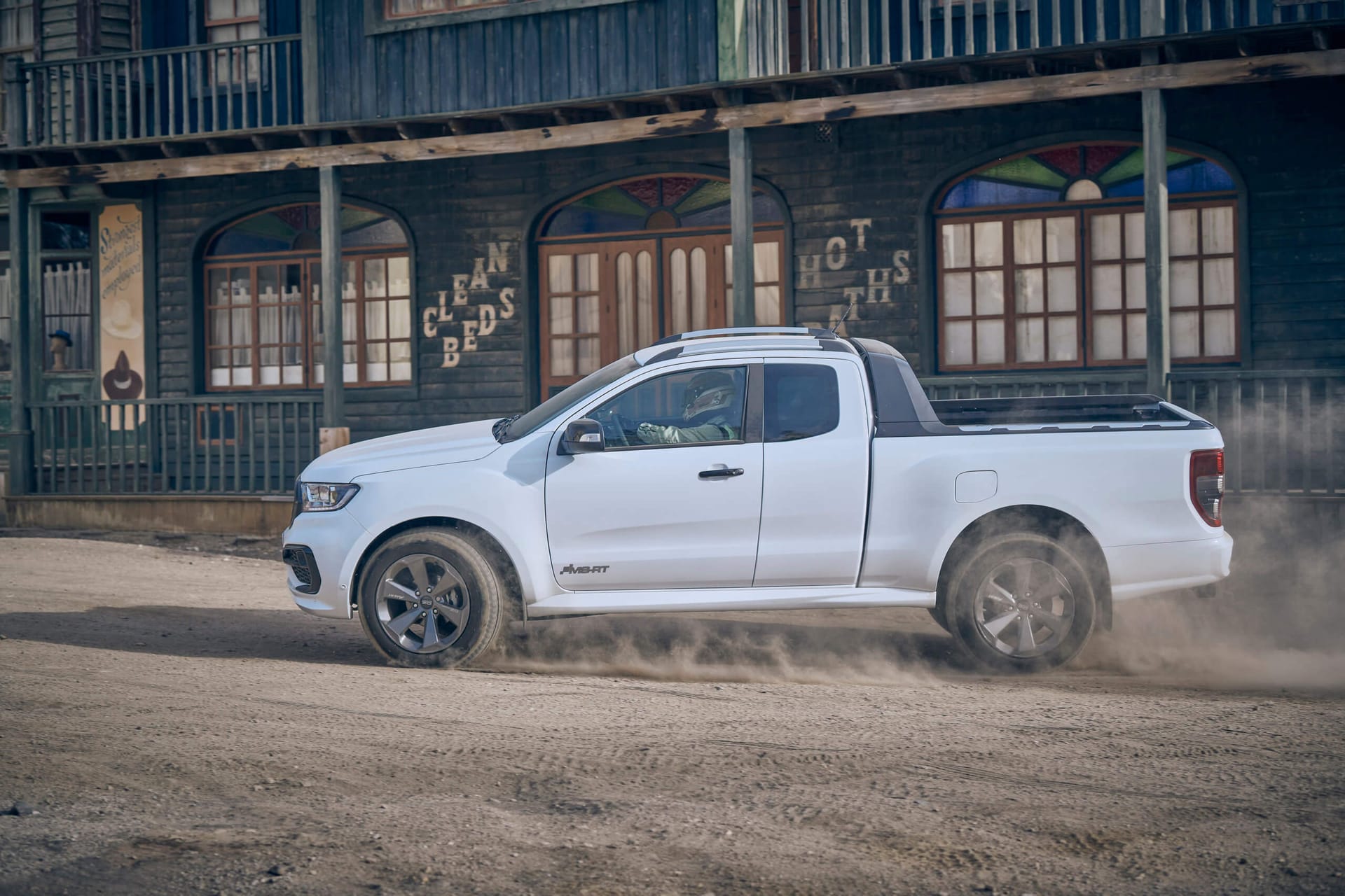Biały Ford Ranger jedzie po zakurzonej drodze przed drewnianym budynkiem z łukowatymi oknami.