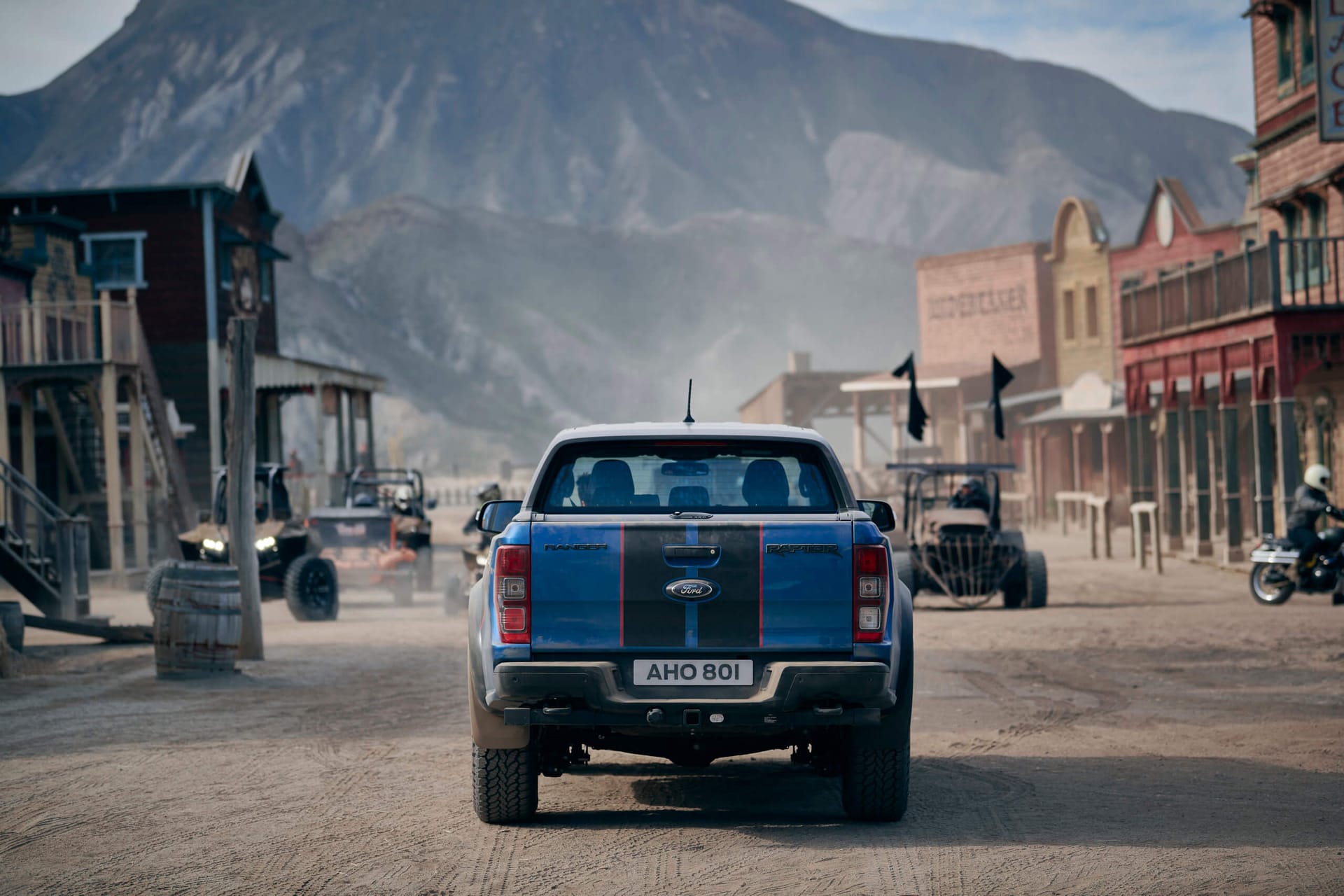 Niebieski Ford Ranger przejeżdża przez zakurzone miasteczko na tle gór i samochodów terenowych.