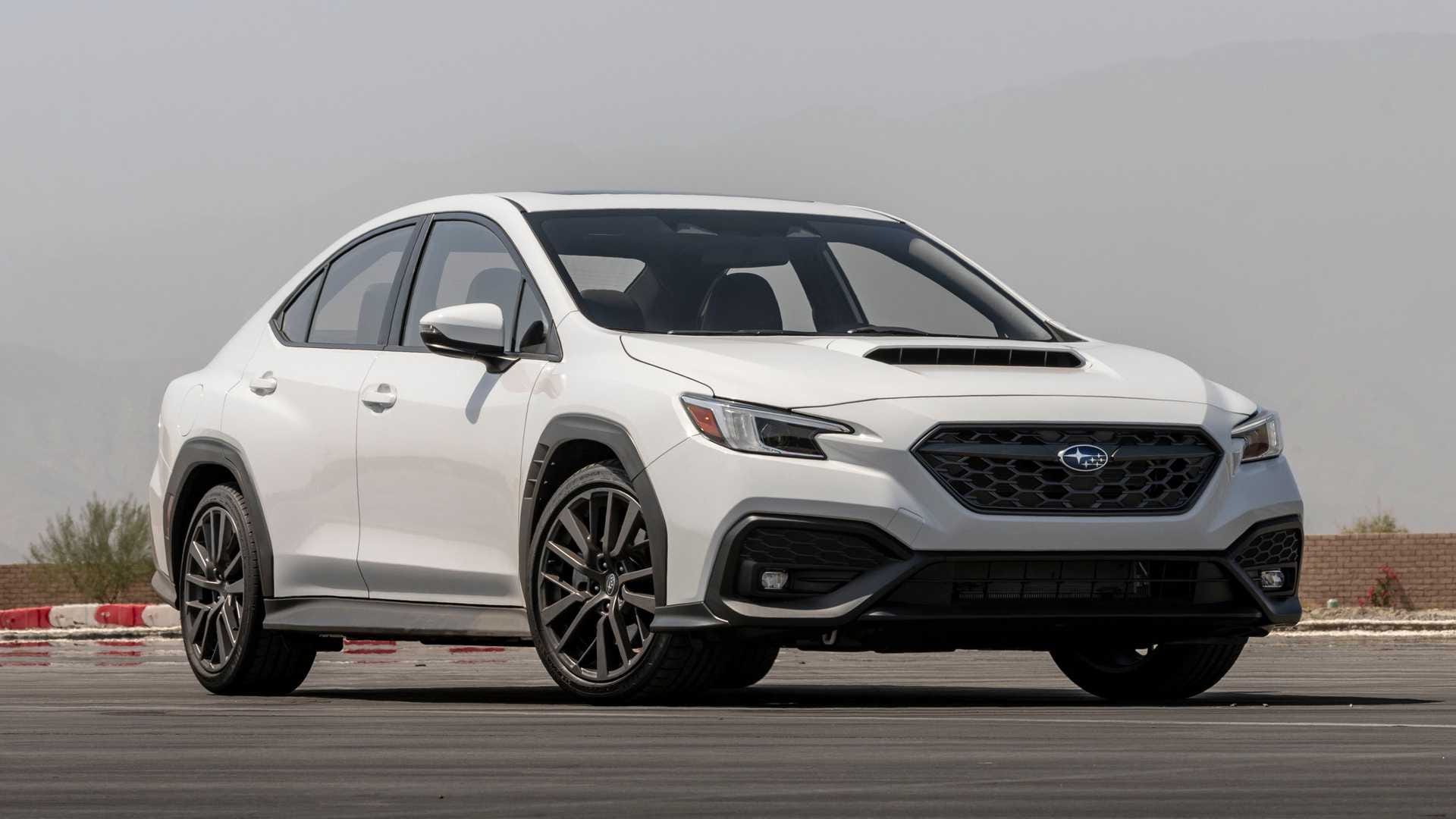 Białe Subaru WRX stoi na asfalcie, a w tle widać góry i ceglany mur pod zamglonym niebem.