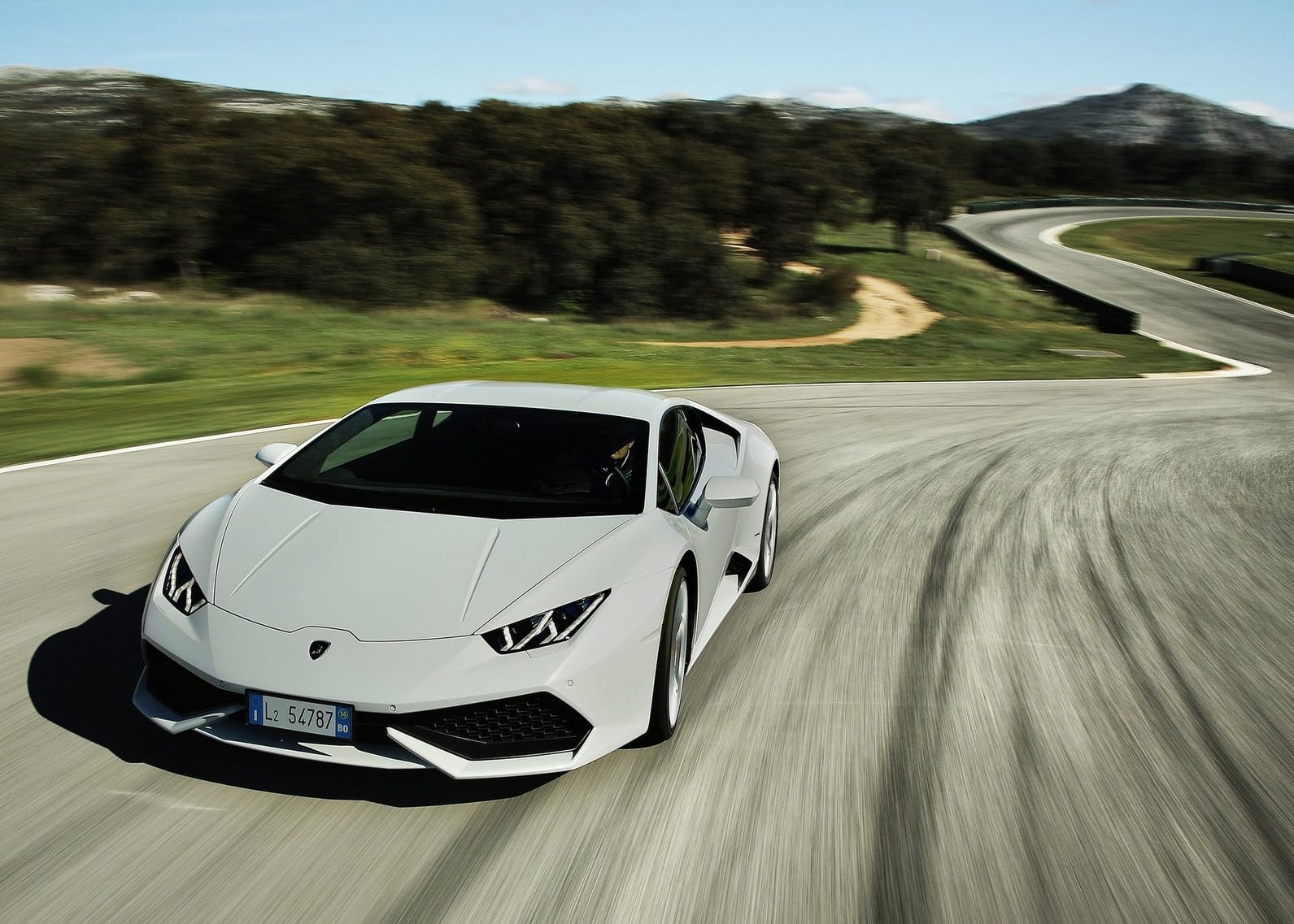 Biały Lamborghini Huracan pokonuje zakręt toru wyścigowego otoczonego drzewami i wzgórzami.