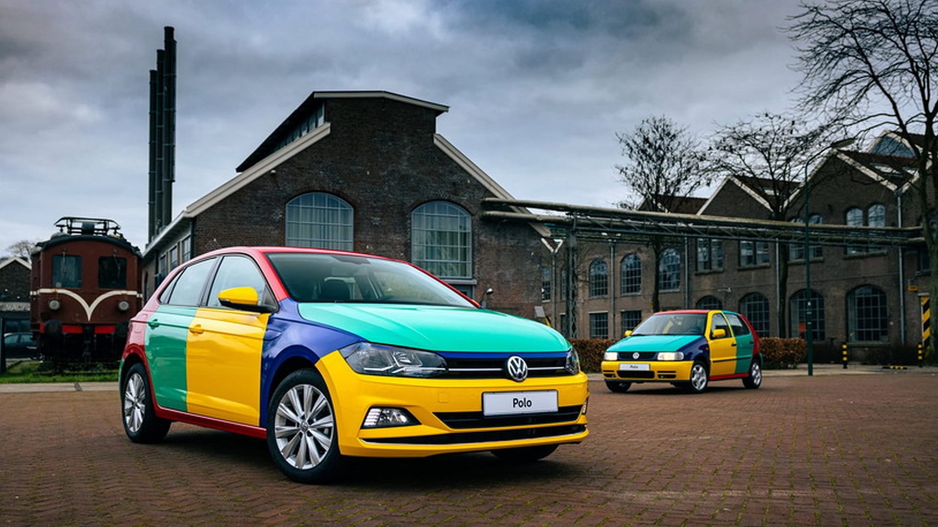 Dwa wielobarwne Volkswageny Polo stoją na kostce brukowej przed budynkami przemysłowymi pod chmurami.