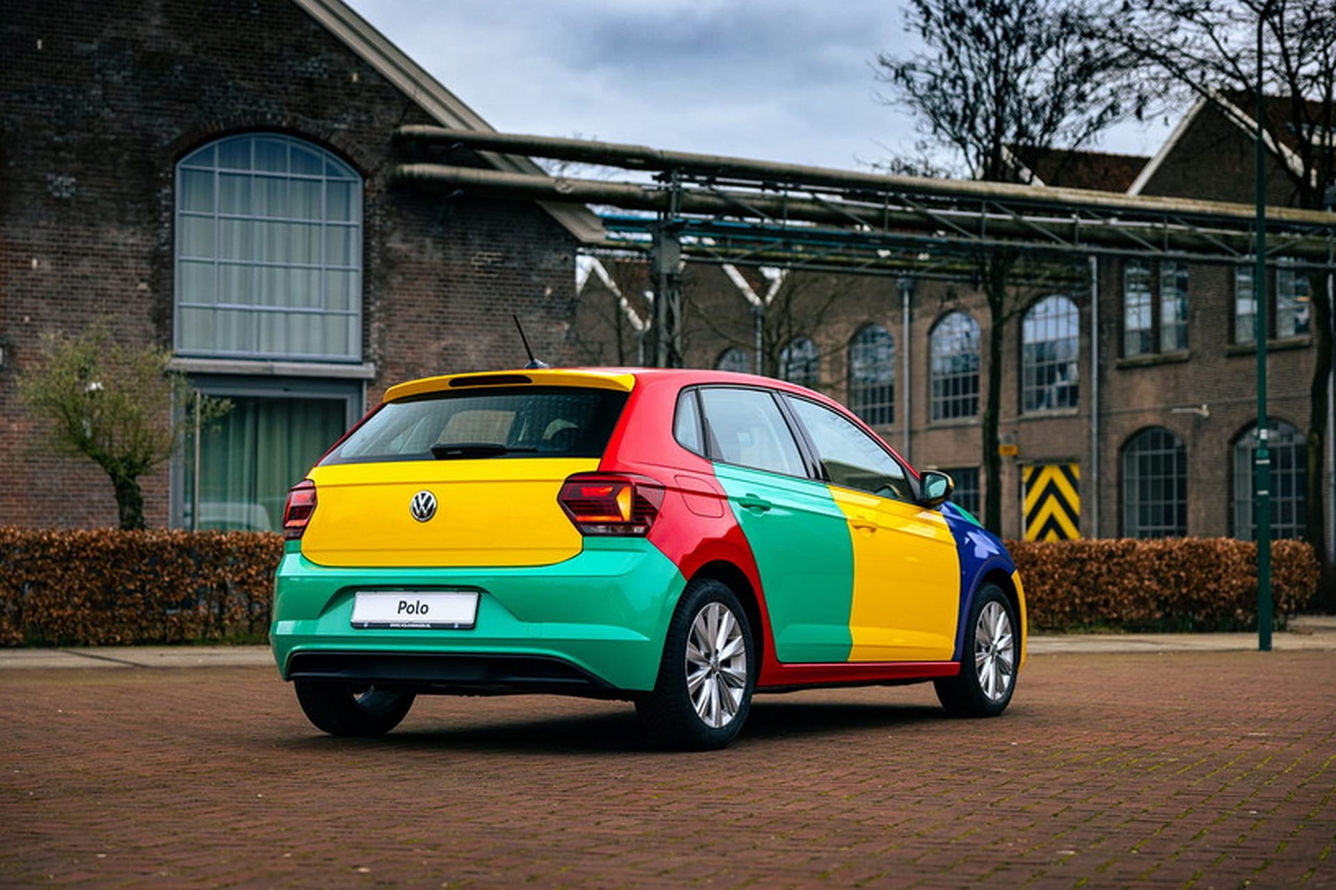 Volkswagen Polo w kolorowym, patchworkowym lakierze stoi przy ceglanych budynkach i drzewach.