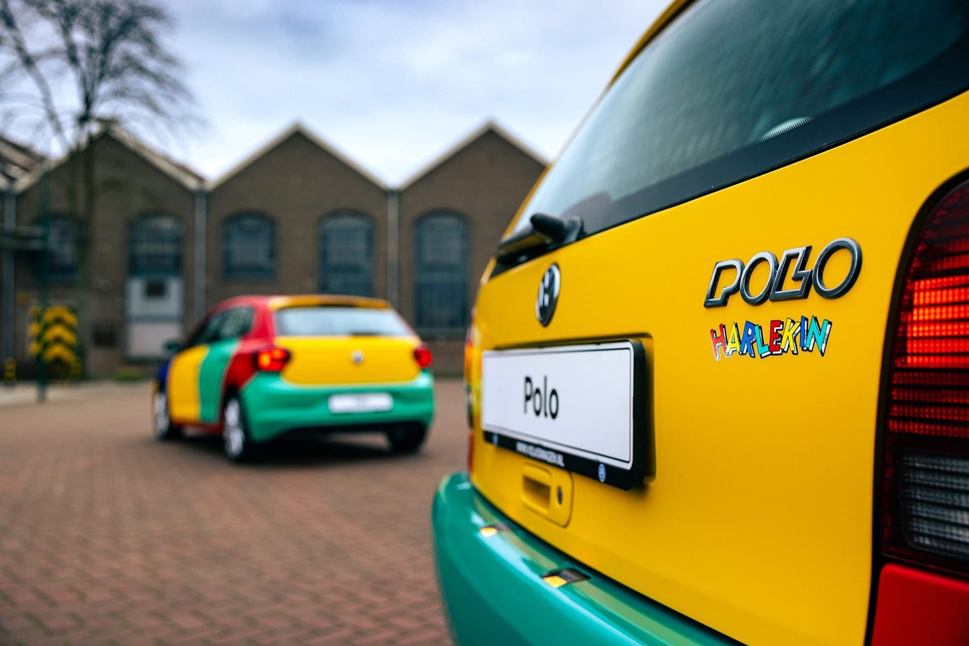Dwa kolorowe Volkswageny Polo Harlekin stoją na brukowanej drodze obok budynków przemysłowych.