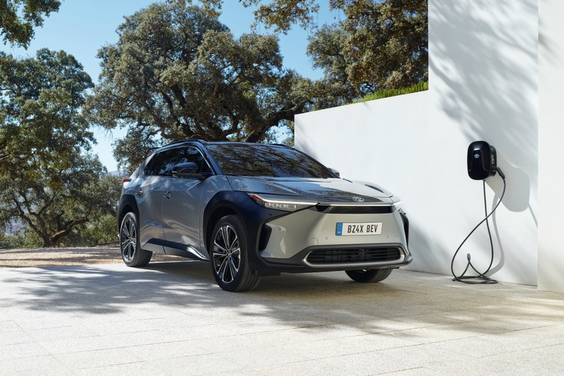 Szara Toyota bZ4X ładuje się przy białej stacji na tle drzew.