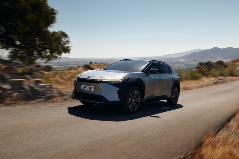 Srebrna Toyota bZ4X jedzie wiejską drogą wśród drzew, a w tle widać góry i czyste niebo.
