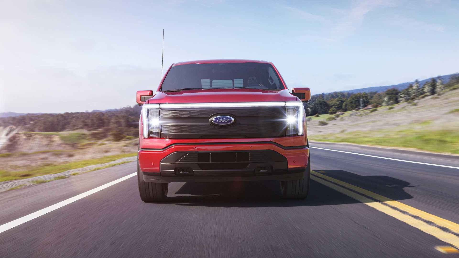 Czerwony Ford F-150 Lightning jedzie dwupasmową wiejską drogą w ciągu dnia – widok z przodu.