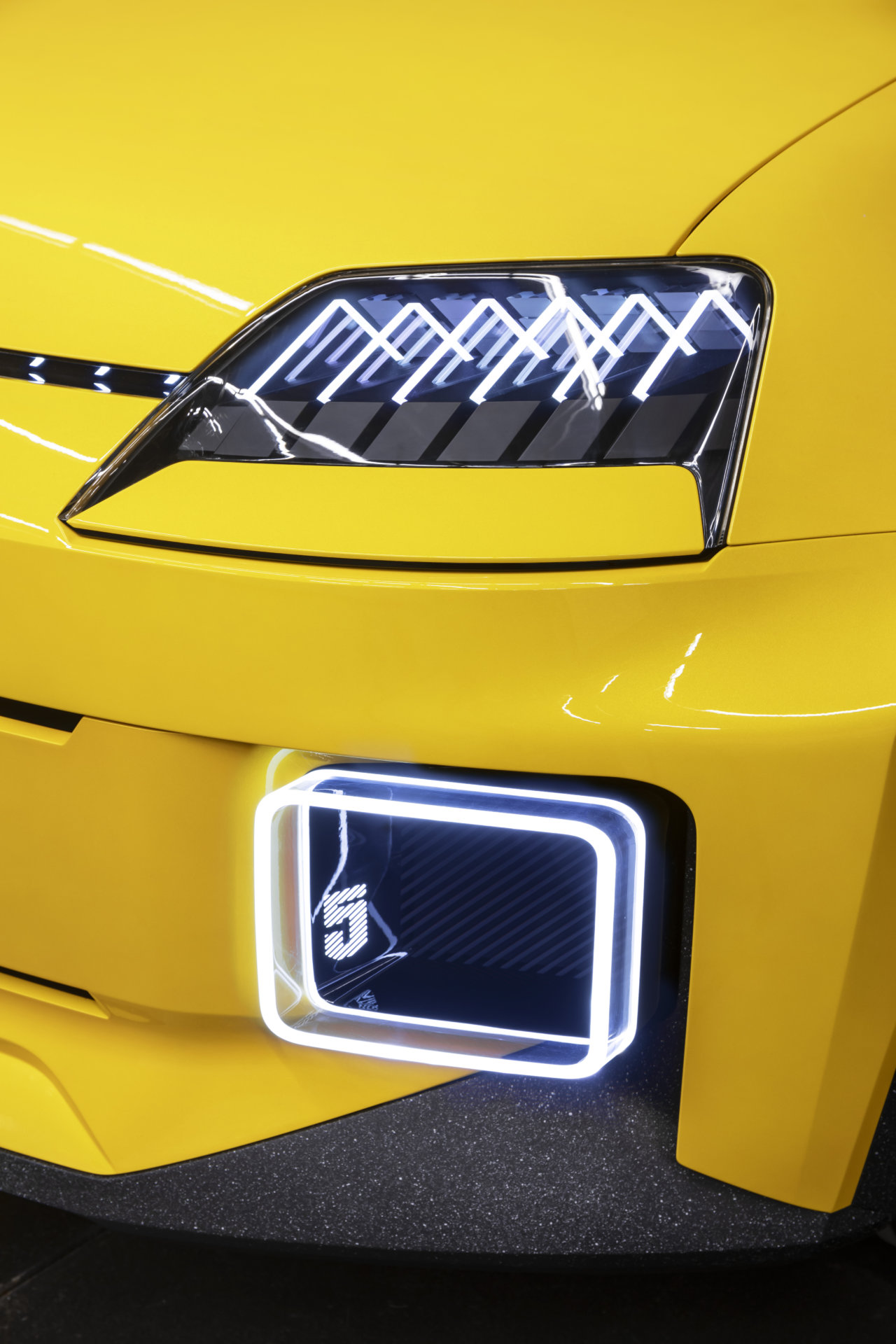 Lewy przedni róg żółtego Renault 5 z reflektorem, świecącą lampą i fragmentem zderzaka.
