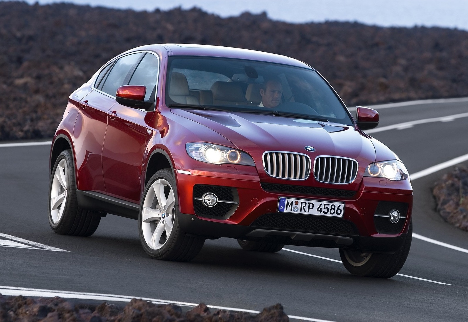 Czerwone BMW X6 jedzie krętą drogą w skalistym terenie; widać kierowcę.