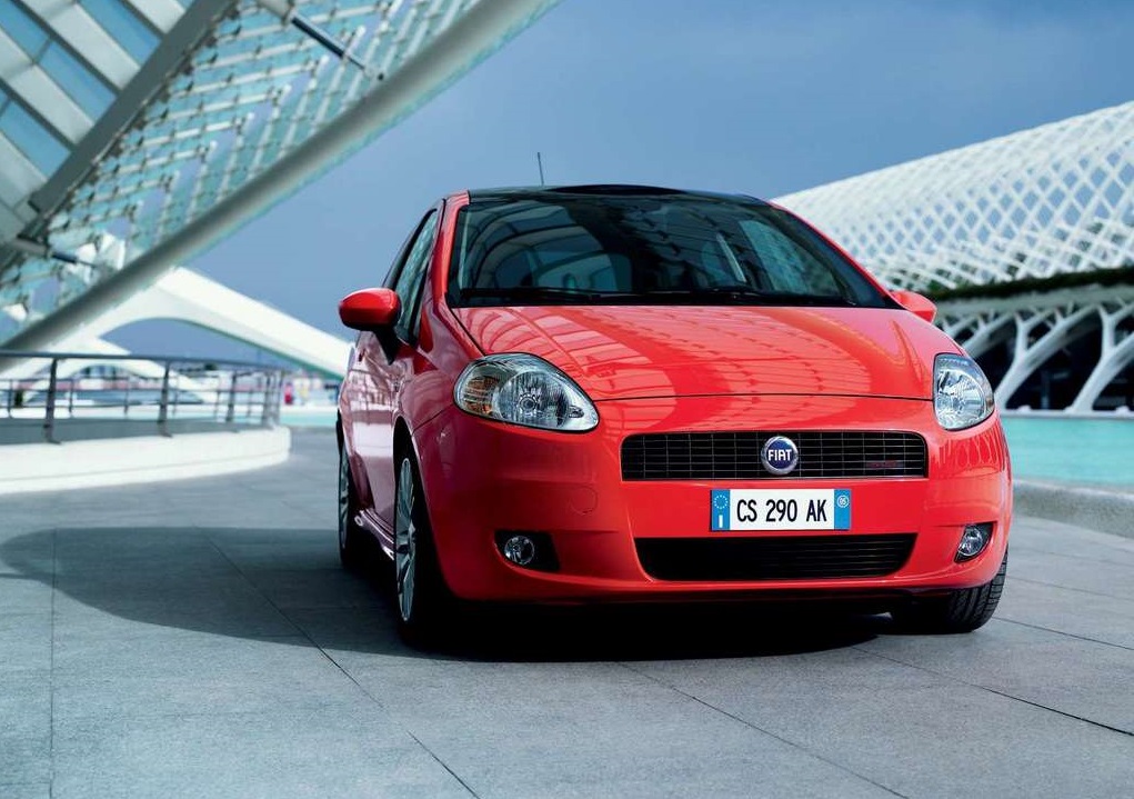 Czerwony Fiat Punto stoi przy białym, geometrycznym budynku w nowoczesnym miejskim otoczeniu.