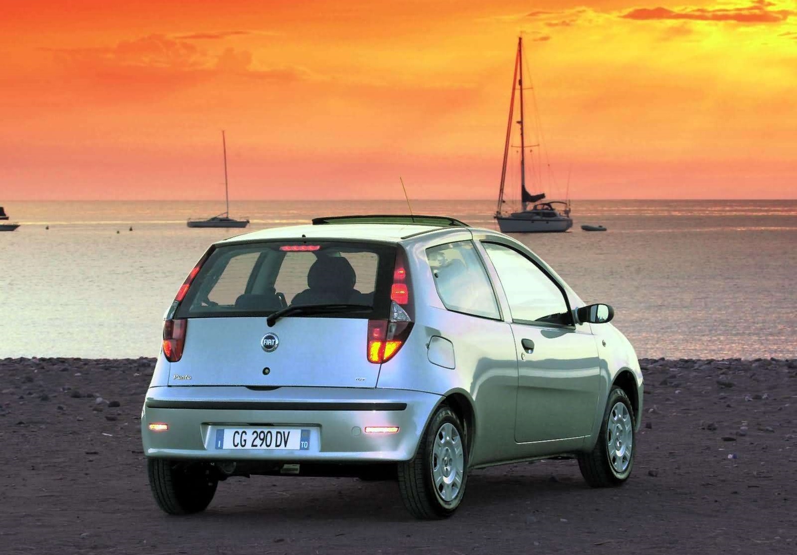 Srebrny Fiat Punto zaparkowany przy plaży o zachodzie słońca, a w tle żaglówki.